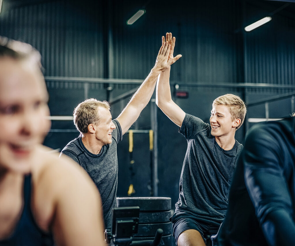 Smiling male friends cycling at health club. Sporty men are high-fiving while working out. They are in cross training gym.